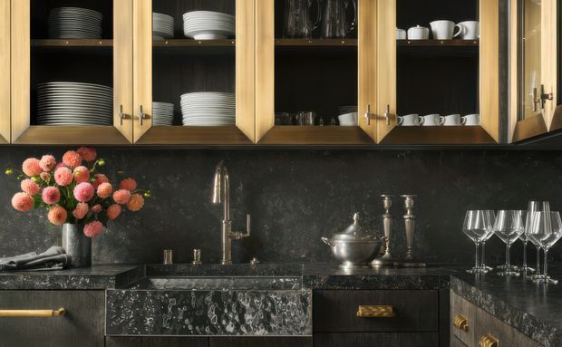 Close-up of an elegant, dark-toned kitchen and drinks station with open timber-framed glass-front cabinetry displaying plates, bowls, cups and glassware. A dark stone backsplash and matching benchtops surround a polished metal sink and tap, with decorative serving pieces and flowers adding warmth. The cabinetry combines light timber doors with dark interior joinery and textured drawer fronts.