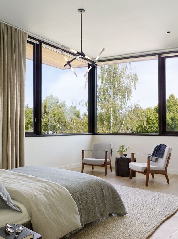Bright, serene bedroom with a large corner of black-framed windows overlooking trees. The room contains a neatly made bed with layered neutral bedding, two mid-century wooden armchairs, a small dark side table with a plant, full-height beige curtains, pale timber flooring, and a textured light rug. A sculptural black-and-white pendant light hangs from the ceiling.