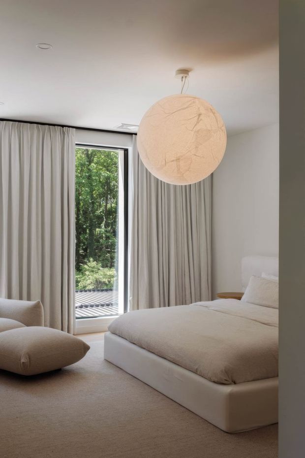 A serene, softly lit bedroom in a neutral palette, featuring a low upholstered bed, plush bedding, a large rounded lounge chair, full-height pleated curtains, and a tall glazed opening overlooking greenery. A large textured spherical pendant light is suspended near the ceiling.