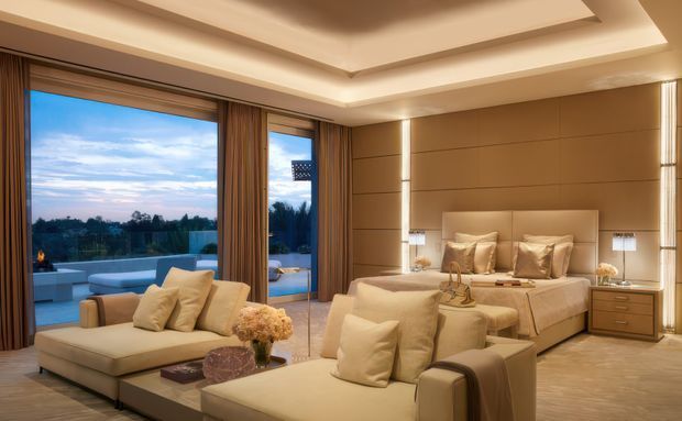A warm, luxurious bedroom suite with a built-in upholstered bed, matching neutral-toned seating, bedside tables, and a layered recessed ceiling. Full-height glazed doors open to an outdoor terrace and overlook a landscaped setting, while timber-toned wall panelling and integrated lighting create a refined contemporary atmosphere.