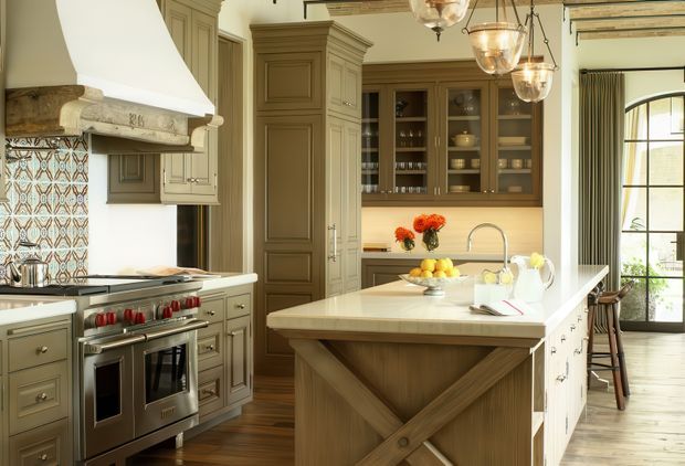 A refined traditional kitchen with a large central island, detailed taupe cabinetry, a professional stainless-steel range, patterned tile backsplash, and glass-front upper cabinets. The island has a light stone benchtop and decorative timber cross-brace detailing. A sculptural white rangehood, pendant lights, timber flooring, and tall black-framed glazing complete the warm, classic interior.