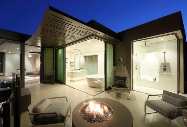 Night-time architectural photograph of a sophisticated outdoor courtyard or terrace centered on a circular fire pit and lounge seating. The space is framed by dark angular roof forms and extensive glazed openings, with a brightly illuminated bathroom featuring a freestanding bathtub and shower visible inside, and a bedroom visible through glazing at left. The courtyard has pale tiled flooring, a slatted soffit, dark-framed doors, and a suspended lounge chair, creating a dramatic indoor-outdoor connection.