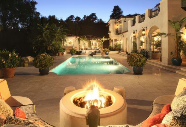 Luxurious Spanish- or Mediterranean-inspired outdoor courtyard at dusk, centred on a rectangular illuminated swimming pool and a circular fire pit in the foreground. Stucco buildings with arched openings, tiled roof sections, balconies, decorative wall lanterns, climbing greenery, potted plants and upholstered lounge seating surround the paved terrace.