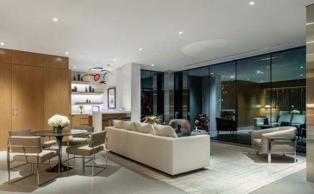 A spacious, open-plan contemporary living and dining area with a cream sectional sofa, glass coffee table, dining table and upholstered chairs. Floor-to-ceiling dark-framed glazing lines the rear wall, while warm timber wall panelling and built-in display shelving add contrast. Recessed ceiling lighting and a large curved ceiling detail create a polished, gallery-like atmosphere.
