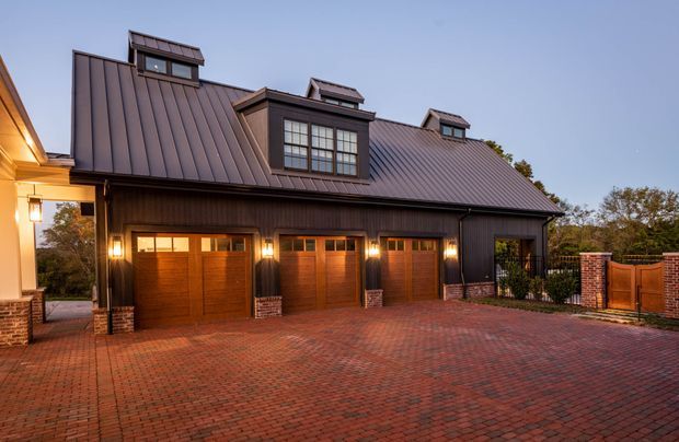 Exterior view of a substantial detached garage or carriage-house-style structure with a dark standing-seam metal roof, multiple dormer windows, dark vertical cladding, and three timber-toned garage doors. The building sits beside a large red brick-paved driveway, with brick piers and low brick walls, warm exterior lighting, and a fenced garden edge. The architecture has a formal traditional character with a barn-like roof form.