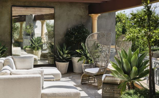 A sunlit outdoor lounge terrace furnished with cream upholstered seating and woven wicker chairs, surrounded by lush potted plants. A large wall mirror reflects the seating and classical columns, while a timber-lined ceiling, stucco walls, and decorative metal balustrade create a relaxed Mediterranean-inspired setting.