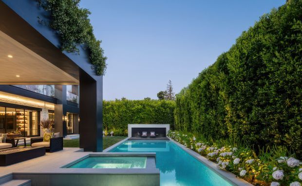 A contemporary residential outdoor entertaining area viewed from a covered terrace, featuring a long rectangular swimming pool with an integrated spa in the foreground, lush clipped hedges, flowering garden beds, and a dark modern pavilion with expansive glazing and warm interior lighting. The home has strong geometric forms, a cantilevered roof, dark structural columns, and timber-toned soffits.
