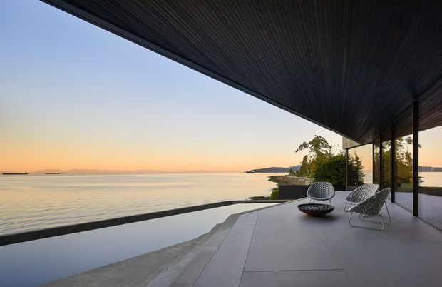 A minimalist waterfront terrace overlooking calm water at sunset. The covered outdoor space features a dark timber-lined soffit, slim dark window or door frames, broad floor-to-ceiling glazing, large pale paving slabs, and two woven lounge chairs with a low fire bowl or table. A reflecting pool or water feature runs along the terrace edge, creating a seamless connection to the coastal setting.