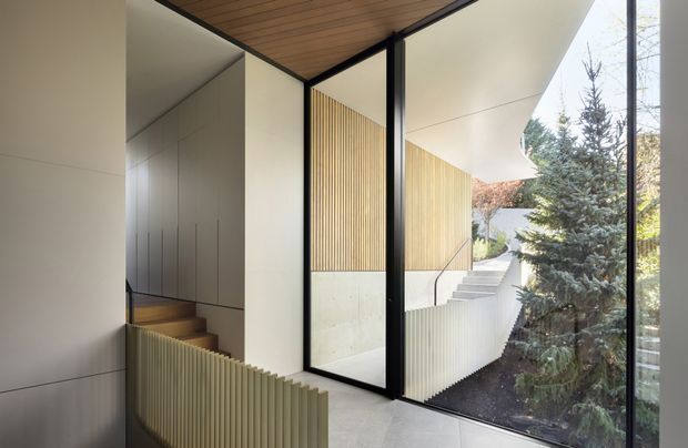 A refined contemporary interior circulation space with a stairway and landing beside full-height black-framed glazing. Warm timber slatted ceiling and vertical timber screening contrast with pale smooth wall panels, concrete-toned stair and floor surfaces, and an adjoining landscaped exterior visible through the glass. The composition emphasizes clean geometric lines, integrated detailing, and natural light.