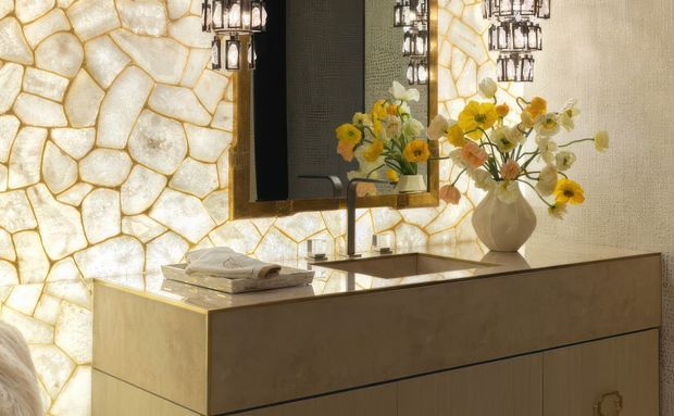 Close-up of a luxurious bathroom vanity or dressing-room console with a large floating-style rectangular mirror, integrated basin and slim faucet, and warm metallic cabinetry. The wall behind is clad in illuminated irregular translucent stone panels with golden veining, creating a dramatic backlit feature. Decorative pendant lights and flower arrangements are reflected in the mirror and placed on the counter.