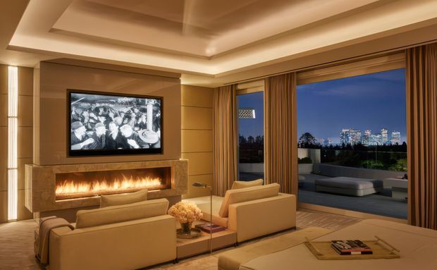 Luxurious contemporary living room with a built-in linear fireplace and television feature wall, multiple tan upholstered sofas and chairs, layered tray ceiling, warm recessed lighting, full-height curtains, and large glazed doors opening to an outdoor terrace with city views. The room has a refined, warm hotel-like atmosphere and is arranged for media viewing and entertaining.