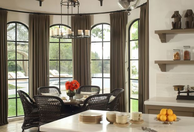 A refined dining area connected to a kitchen, featuring a round dining table with woven dark chairs, a large traditional-style chandelier, and tall arched black-framed windows dressed with full-length taupe curtains. A light kitchen island with stacked plates, cups, cutlery, and a bowl of citrus occupies the foreground. Built-in shelving and a pale stone or plaster backsplash are visible on the right. The room has a warm, elegant, classic character with abundant natural light.