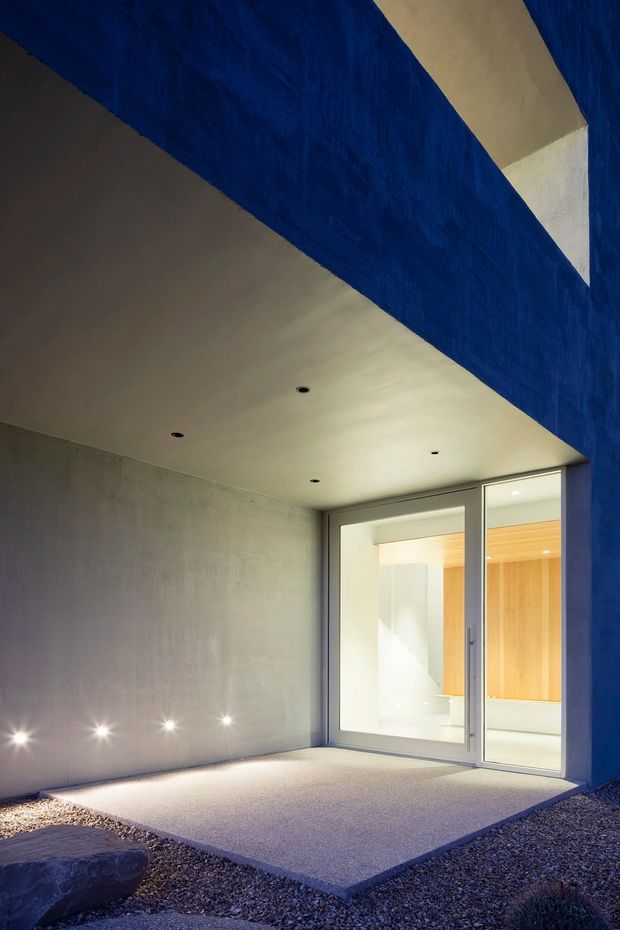 A minimalist architectural exterior entry court beneath a dramatic deep-blue sculptural overhang. A smooth concrete wall and ceiling frame a large glazed door opening into a brightly lit interior with warm timber wall lining. Recessed downlights illuminate the covered concrete threshold, surrounded by dark gravel and a large natural stone.