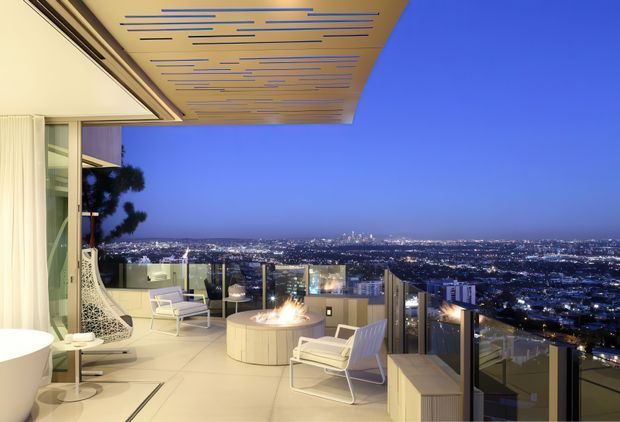 A luxurious elevated outdoor terrace at dusk, overlooking a sprawling illuminated cityscape. The terrace features pale tiled flooring, glass balustrades, white contemporary lounge chairs, a sculptural hanging chair, built-in pale cabinetry or seating, and a circular fire feature. A large cantilevered ceiling with linear timber-toned detailing provides shelter, while the adjacent interior is visible at the left edge.