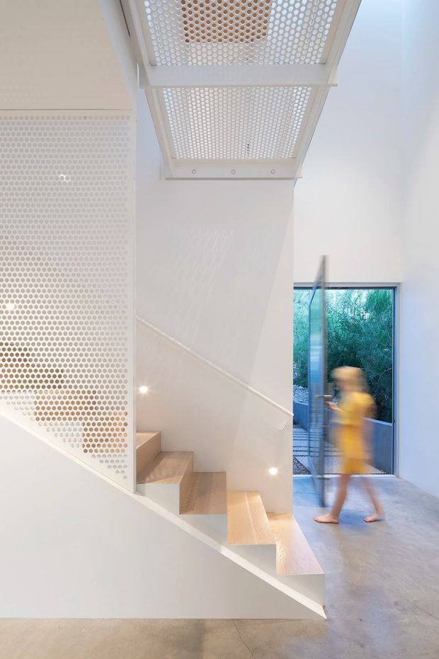 Bright minimalist interior showing a sculptural stairway with pale timber treads, white walls, and a perforated white privacy screen. A large roof or ceiling skylight with a circular perforated grille casts patterned light into the stair hall. A glazed doorway opens to greenery outside, and a blurred person is walking through the space. The floor appears to be smooth polished concrete.