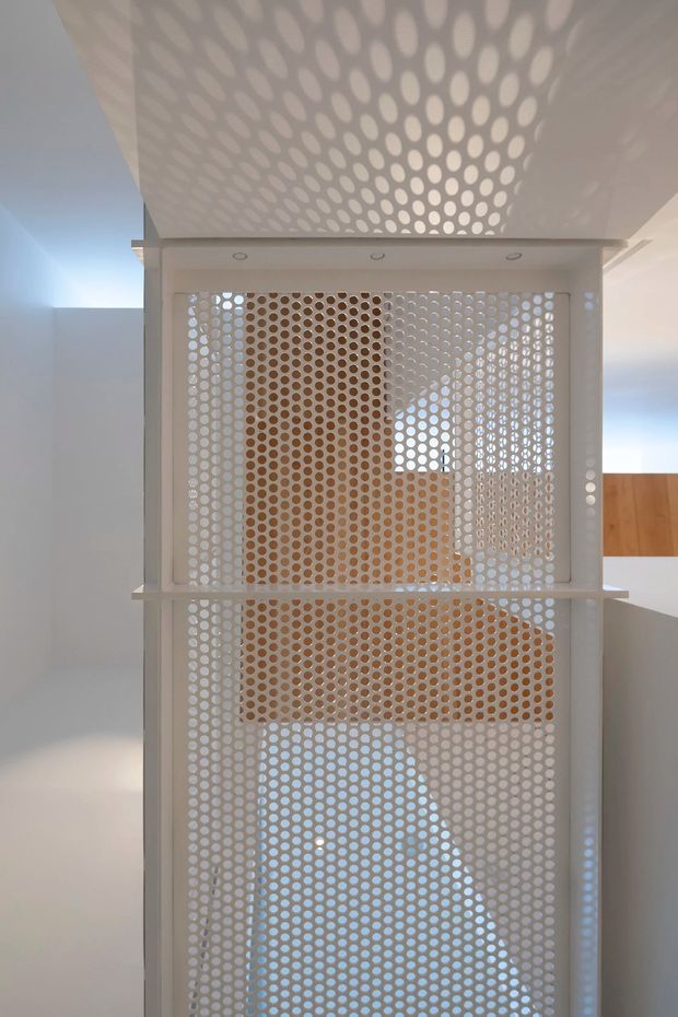 Interior architectural detail showing a tall white perforated screen or balustrade with repeating circular openings. The screen spans multiple levels and casts a distinctive dotted pattern across the ceiling, while glimpses of timber-toned surfaces and bright adjacent areas are visible behind it. The image focuses on the perforated architectural feature rather than a clearly identifiable room.