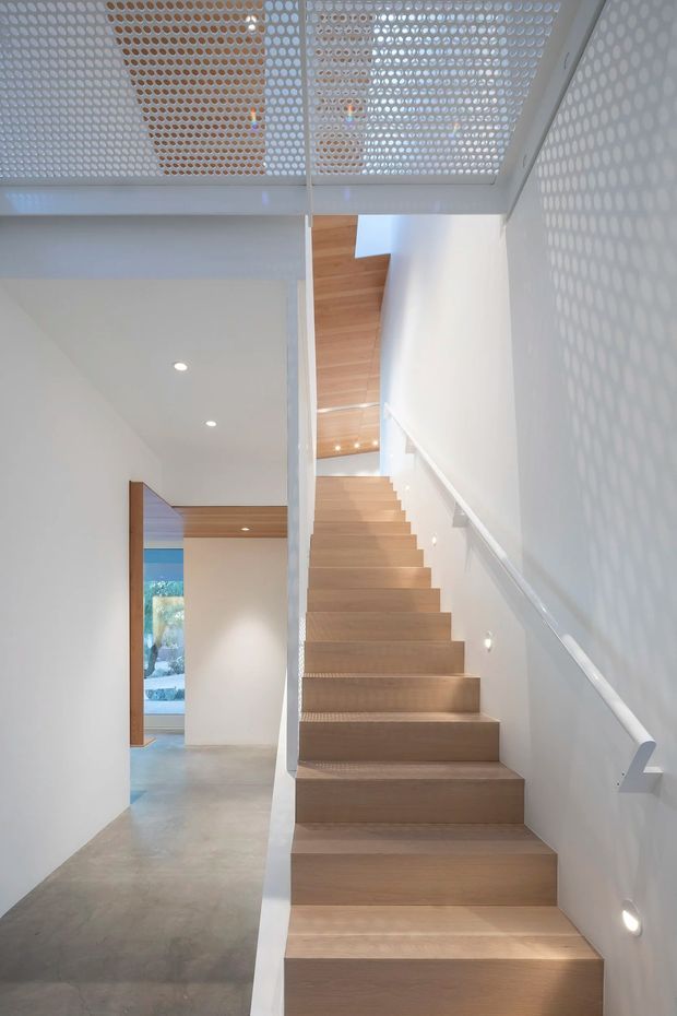 A bright contemporary interior hallway featuring a straight timber staircase with minimalist white handrails and walls. The stair rises beneath a perforated white ceiling screen that casts a dotted pattern of light and shadow onto the adjacent wall. Polished concrete flooring, recessed downlights, warm timber ceiling and door detailing, and a glimpse toward an exterior garden create a clean, airy architectural composition.