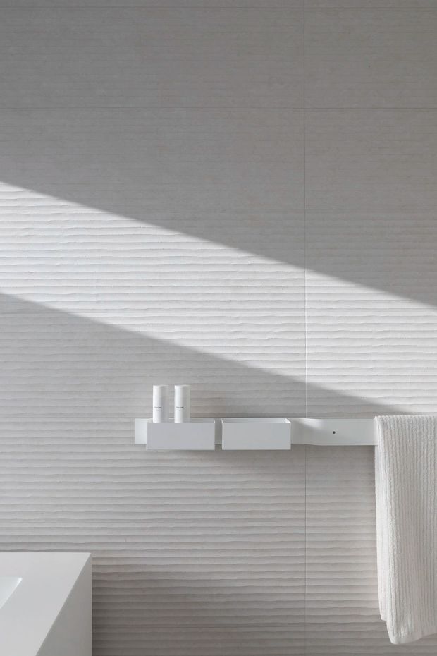 Close-up minimalist interior detail showing a pale grey, horizontally ribbed wall surface with a strong diagonal beam of sunlight. A simple white floating shelf or ledge holds two cylindrical bottles, with a white towel hanging at right and part of a white fixture or counter visible at lower left. The composition is monochromatic, tactile, and highly pared back.
