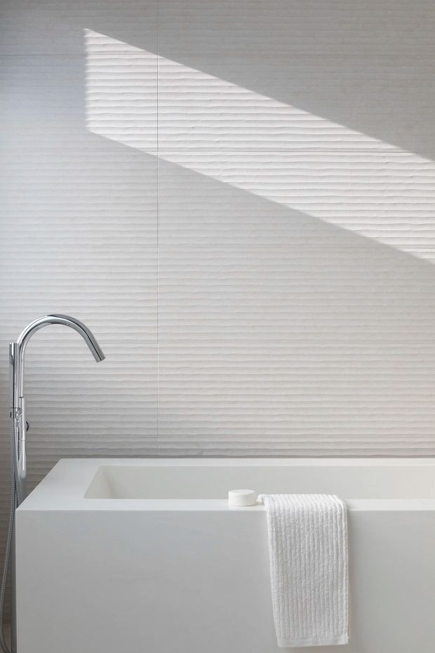 Minimalist bathroom featuring a freestanding white bathtub, chrome floor-mounted tub filler and hand shower, and a textured white wall tile backdrop. Strong diagonal sunlight and shadow create a sculptural, serene atmosphere, with a neatly draped white towel adding softness.