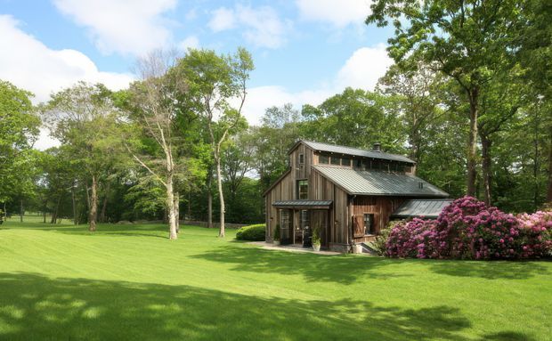 A rustic timber-clad barn-like residence or outbuilding set within a spacious, manicured green lawn and mature woodland. The structure has a steep gabled roof, a long clerestory-like upper window band, dark metal roofing, timber-framed glazing, and a covered entry or porch. Pink flowering shrubs cluster along the right side of the building, while the overall scene is bright and pastoral.