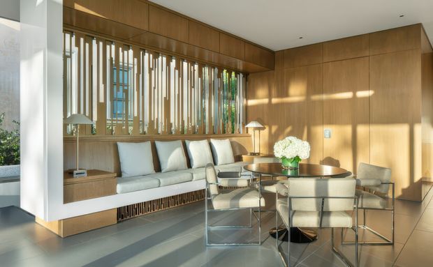 A refined open-plan living and dining space with extensive warm timber wall panelling and built-in joinery. A long built-in window seat with pale cushions sits beneath a decorative vertical timber screen, while a compact round dining table with upholstered metal-framed chairs occupies the foreground. Large glazing brings in strong natural light, and the room has a clean, tailored contemporary character.