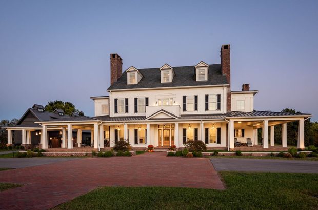 A large symmetrical two-storey white residential house viewed from the front at dusk. The home has a traditional formal facade with black shutters, tall multi-pane windows, brick chimneys, a dark shingle roof with dormer windows, a central entrance with pediment, and broad covered verandas or porticos supported by white columns. A brick-paved driveway and landscaped lawn occupy the foreground.