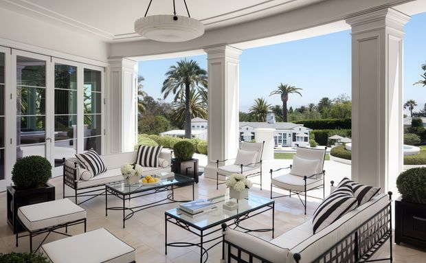 Bright, elegant covered outdoor living veranda with open sides, classical white columns and detailed ceiling mouldings. The space is furnished with white cushioned sofas and lounge chairs featuring black-and-white striped accents, glass-top metal coffee tables, planters, and decorative lighting. It overlooks a landscaped residential setting with palm trees and a pool visible beyond.