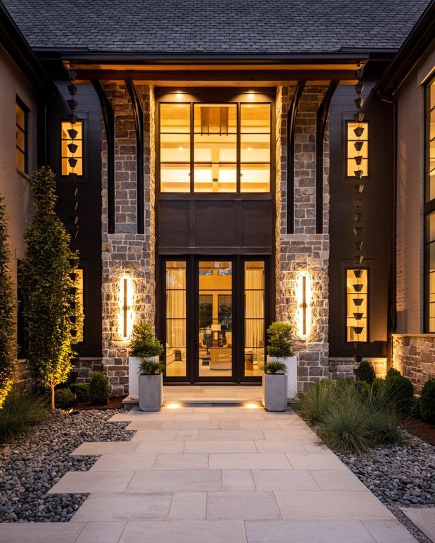 A dramatic dusk view of a grand residential entrance. The facade features dark cladding, substantial rough-cut stone piers, a projecting timber-framed canopy, black-framed glazing and doors, and warm illuminated interior and exterior lighting. A broad pale stone-paved path leads through a landscaped entry court with clipped shrubs, ornamental grasses, gravel beds, and large planters.