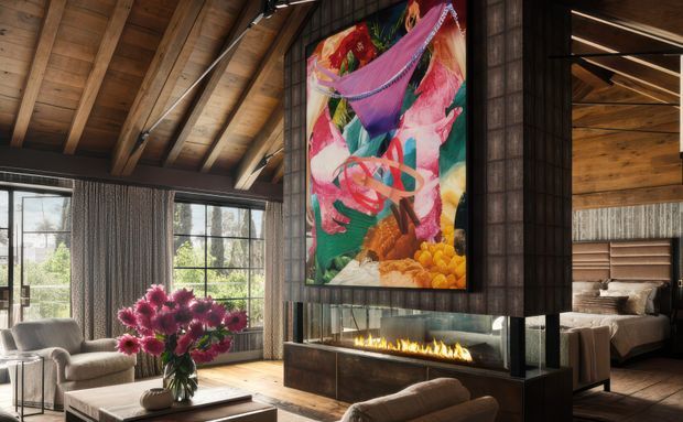 A warm, richly detailed living room with a dramatic exposed timber vaulted ceiling, expansive black-framed windows, and dark vertical timber wall cladding. A large colourful artwork hangs above a long built-in linear fireplace, with a bedroom visible through an opening to the right. Neutral upholstered seating and a vase of pink flowers complete the inviting interior.