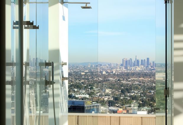 A bright, high-rise glass bathroom with a frameless shower enclosure and square overhead showerhead, overlooking a dense city skyline through floor-to-ceiling glazing. The space features minimal architectural detailing, clear glass, pale surfaces, and a vertical timber-toned screen or wall element at the lower edge.