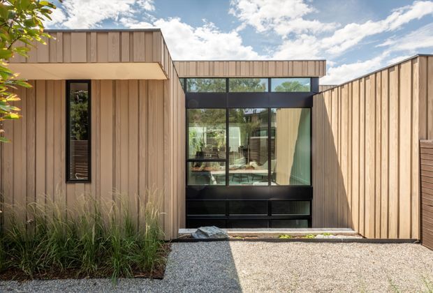 A contemporary single-storey residential facade viewed from outdoors, composed of warm vertical timber cladding and strong black-framed glazing. The central glazed link reveals an interior living or dining area, while planted grasses and a pale gravel forecourt soften the minimalist geometric composition. The building has low horizontal roof forms and a sheltered recessed entry or connection between timber-clad volumes.
