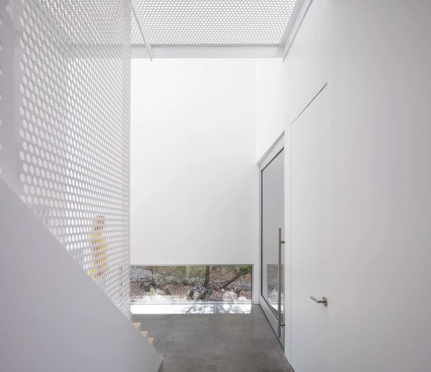 Minimalist white stairway and landing with a perforated screen balustrade and matching perforated overhead roof or skylight panel. A glazed door opens toward a small landscaped courtyard, while the polished grey concrete floor and seamless white walls create a bright, restrained architectural interior.