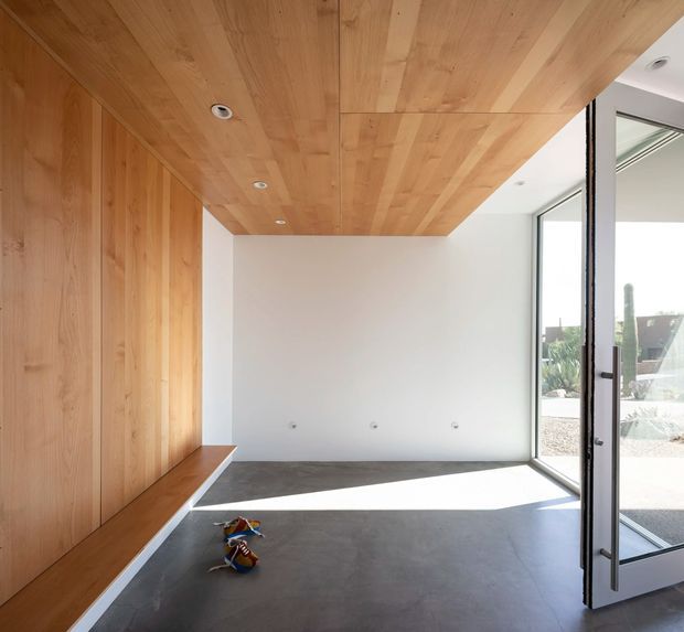 Bright, minimalist interior room with a full-height timber-lined wall and matching timber soffit ceiling. The space has a smooth dark grey concrete-like floor, plain white walls, recessed downlights, and large black-framed glazed doors opening to an exterior courtyard or garden. Built-in timber bench seating runs along the wall, and the room appears largely unfurnished.