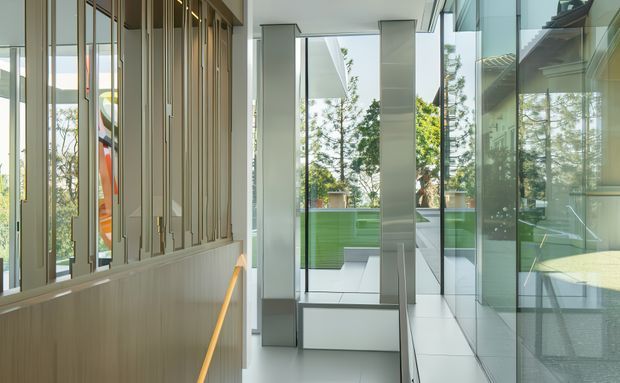 A bright contemporary interior corridor or landing with full-height glazing and reflective glass walls. Vertical timber battens form a feature screen along the left side, while pale tiled flooring, slim grey structural columns, and extensive windows create a light-filled connection to an exterior terrace or courtyard. A vivid green built-in exterior wall or screen is visible beyond the glazing.