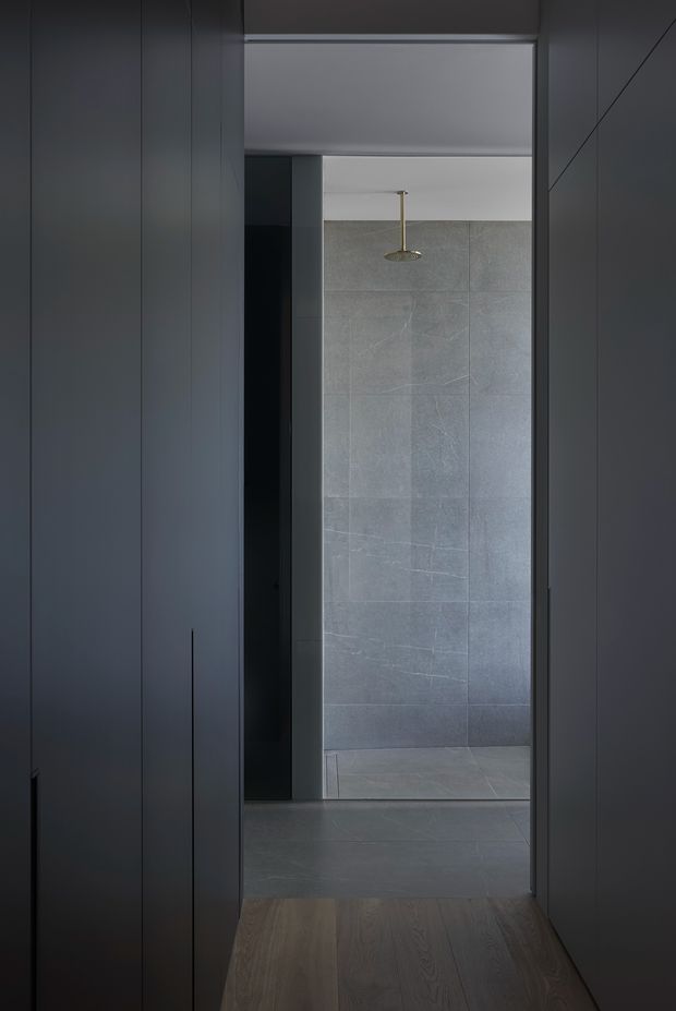 A narrow, moody view through a dark built-in passage into a minimalist bathroom. The bathroom features large grey stone-look wall and floor tiles, a walk-in shower with a slim brass ceiling-mounted shower fixture, and dark flush cabinetry or wall panelling framing the opening. A warm timber floor leads toward the cool grey tiled space.