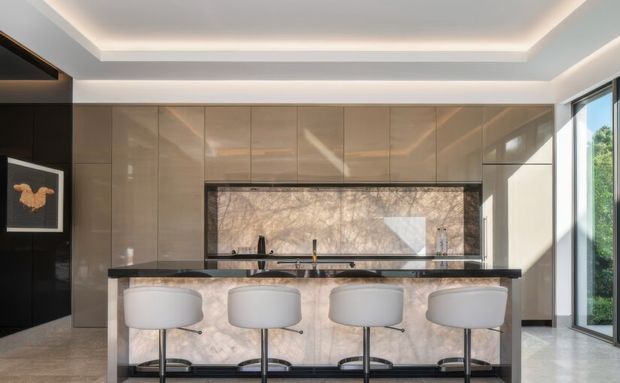 A refined contemporary kitchen with a long island and four white bar stools. Glossy taupe-beige full-height cabinetry spans the rear wall, with a dark stone island benchtop and a large marble-look splashback beneath upper cabinetry. Integrated warm LED cove lighting outlines the recessed ceiling, while full-height glazing on the right brings in daylight and greenery. A black feature wall with framed artwork is visible at left.