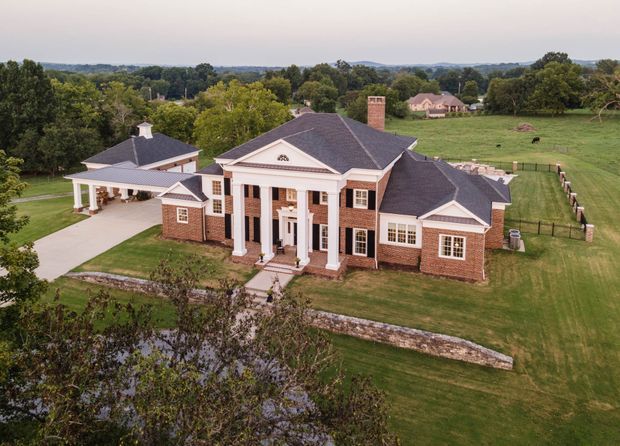 Aerial view of a large, symmetrical red-brick country residence with white classical columns, pediments, multi-pane windows, dark hipped roofs, and a prominent central entrance. The house includes a covered driveway/carport structure and is set within expansive landscaped lawns and fenced rural grounds.