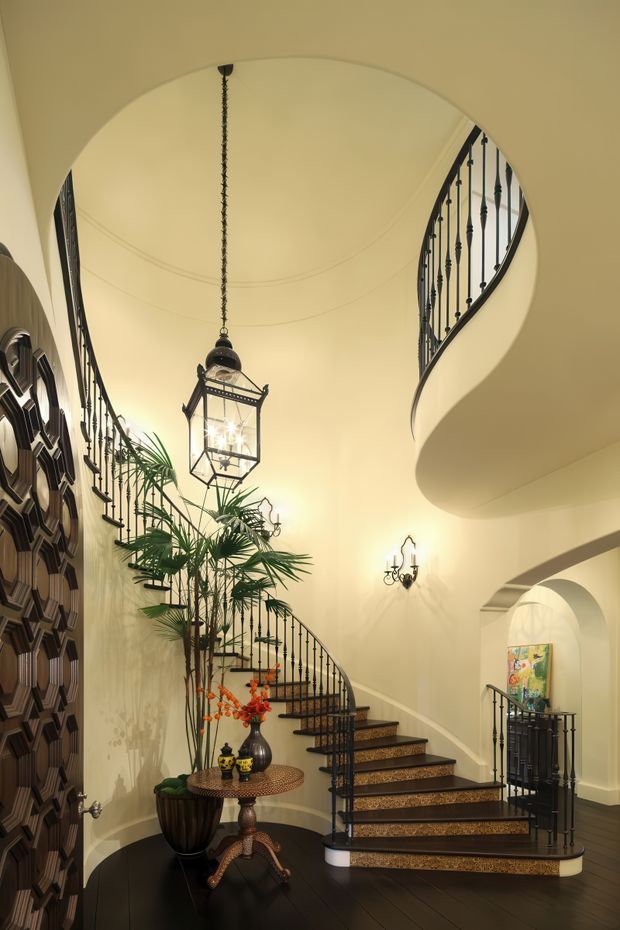 A grand residential foyer with a sweeping curved staircase, ornate black metal balustrades, dark wood flooring, and a high arched ceiling. A large hanging lantern, wall sconces, tropical plants, a round console table, and decorative wall panelling create an elegant, traditional atmosphere.