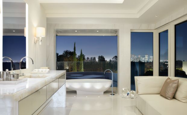 A luxurious contemporary bathroom with a freestanding white bathtub positioned in front of expansive floor-to-ceiling glazing, offering views of trees and a distant illuminated city skyline. The room features a large marble vanity with mirrored cabinetry, twin vessel sinks and polished chrome tapware, a cream built-in sofa, glossy light flooring, and layered recessed ceiling detailing. The palette is predominantly white and cream with blue-toned evening views beyond the glass.