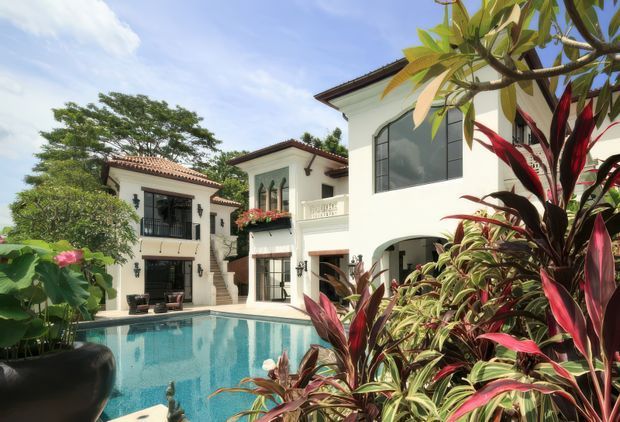 A bright Mediterranean-inspired residential courtyard viewed from beside a swimming pool. The multi-wing white stucco building has terracotta tiled roof edges, dark-framed windows and doors, arched openings, balconies, and decorative exterior lighting. Lush tropical planting surrounds the turquoise pool, creating a resort-like outdoor setting.