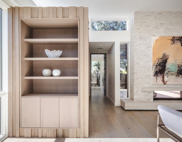 Bright contemporary living room with a custom light timber built-in shelving and cabinet wall, framed by vertically fluted timber detailing. The room features warm timber flooring, a white-painted ceiling and walls, a pale textured brick feature wall, and a minimalist suspended fireplace hearth beneath abstract artwork. A hallway and glazed doorway extend beyond the main space.