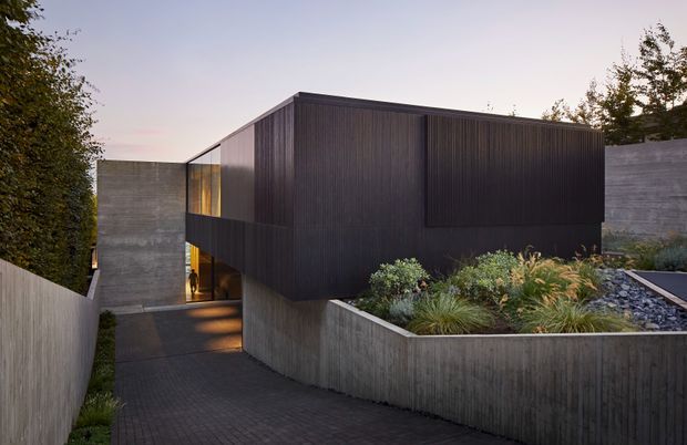 A contemporary dark-clad residential building viewed from an external landscaped approach. The compact, box-like upper volume is clad in vertically ribbed dark material and appears to cantilever over a recessed glazed entry. Raw concrete retaining walls and planter walls frame a gently descending paved walkway, with drought-tolerant planting and dark stone landscaping around the structure. Warm interior light glows through the entry glazing at dusk.