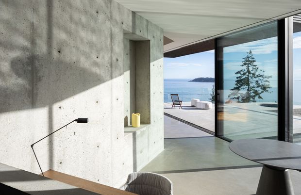 A minimalist concrete interior opening onto a waterfront terrace through large sliding glass doors. The space features exposed raw concrete walls with recessed shelving, a smooth concrete floor, a simple black floor lamp, and the edge of a dining table and chair. The terrace and blue water beyond are visible through the glazing, creating a bright, refined indoor-outdoor composition.