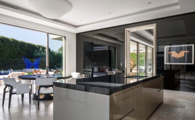 A sophisticated open-plan kitchen and dining area with a large central island featuring dark polished stone benchtops and glossy dark cabinetry. The space has white walls, a recessed tray ceiling with a large circular light fitting, extensive black-framed glazing and mirrored surfaces, and a dining table overlooking a landscaped garden and pool area. The overall design is refined, modern, and high-contrast.