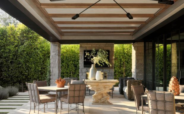 A sophisticated covered outdoor entertaining area with a dining table and chairs, a substantial stone outdoor table or island, and a sculptural fireplace feature at the rear. The space has exposed timber ceiling beams with integrated linear lighting, substantial pale stone columns and walls, large black-framed glazed doors, and views into a dense landscaped garden. The design blends contemporary glazing and lighting with natural stone and timber detailing.
