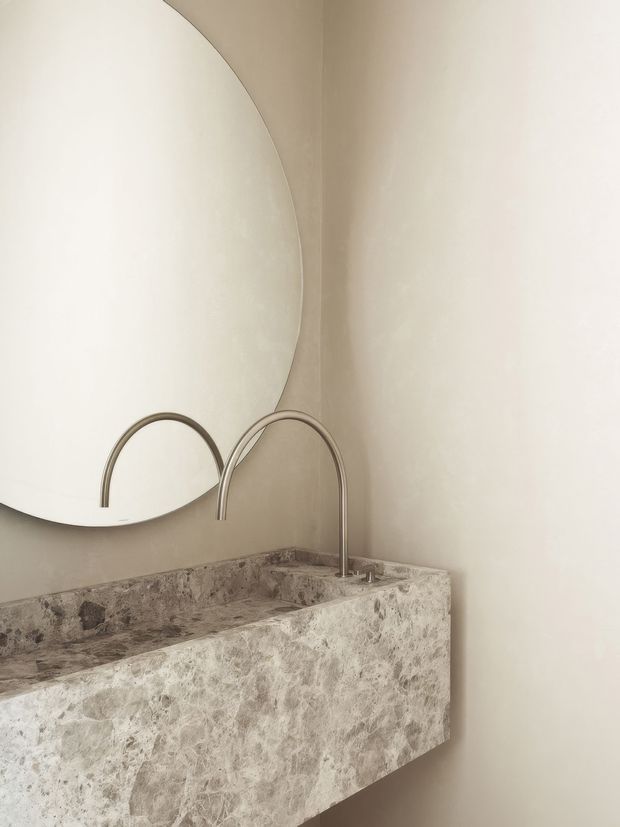Close-up of a minimalist bathroom vanity featuring a floating, solid-stone basin and matching thick stone benchtop with a textured grey-beige natural pattern. Two slim arched metal tap fixtures rise from the basin, while a large round frameless mirror is mounted on the warm off-white plaster wall above. The composition is softly lit, neutral, refined, and sculptural.