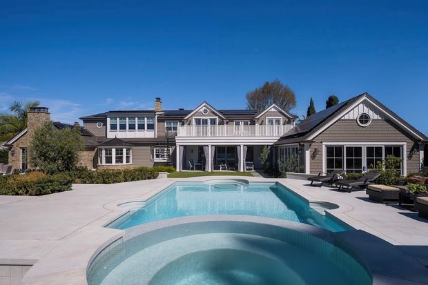 A large multi-wing residential home viewed from a landscaped pool courtyard. The house has a symmetrical, traditional coastal-inspired facade with grey shingle-like cladding, white trim, gabled roofs, dormer-style upper windows, balconies, and a prominent circular gable window. A large pale-blue swimming pool with a curved spa occupies the foreground, surrounded by light paving and outdoor loungers. The scene is sunny, polished, and resort-like.