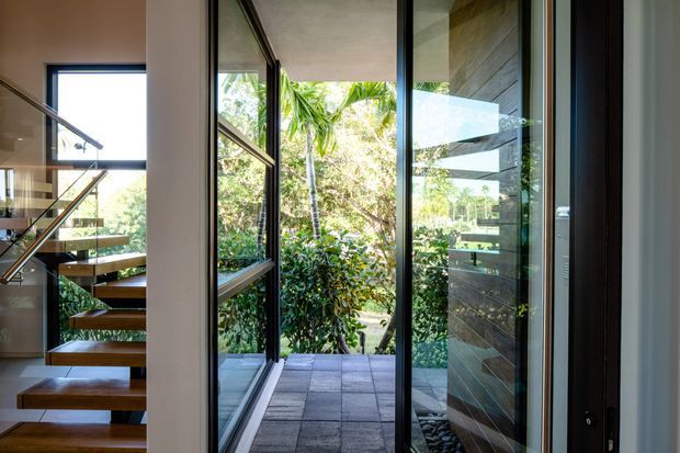 Interior view toward a glazed garden-facing opening, with a floating timber staircase and glass balustrade on the left. Full-height dark-framed glazing and a pivot or hinged glass door lead to a tiled outdoor terrace surrounded by dense tropical planting. A dark horizontal timber or stone feature wall is visible on the right. The scene is bright, contemporary, and strongly connected to the garden.