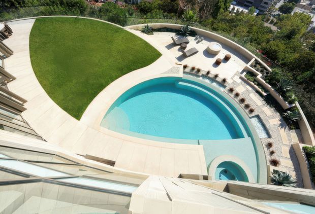 Aerial view of a landscaped residential pool area with a large curved turquoise swimming pool, attached circular spa, pale paved terraces, outdoor loungers, sculptural planters, and a sweeping curved boundary wall. A manicured lawn occupies one side, with the surrounding suburban landscape visible beyond the property.