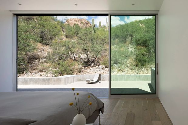 A minimalist bedroom with a low-profile bed in the foreground, pale wood flooring, white walls, recessed ceiling lights, and expansive black-framed floor-to-ceiling glazing. The glazing opens toward a small terrace with a sculptural lounge chair and a rugged desert landscape featuring scrub and cacti. The room is bright, sparse, and strongly connected to the outdoors.